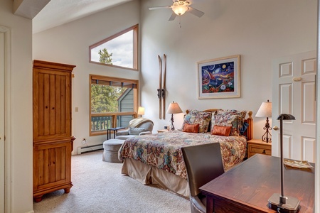 A bedroom with a double bed, colorful bedding, wooden wardrobe, desk, armchair, mounted skis, and large window. Walls are white with an art painting and a slanted ceiling with a fan.