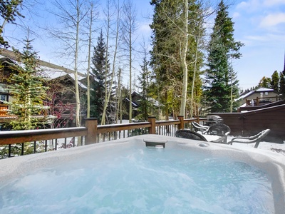 Outdoor hot tub on a snowy deck, surrounded by tall trees and adjacent buildings in a forested area.