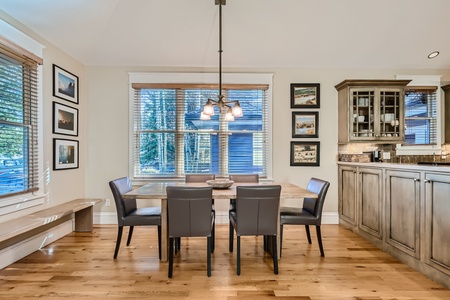 A modern dining area with a wooden table and four gray chairs. Large windows with blinds provide natural light, and framed pictures adorn the walls. A hanging light fixture is above the table.