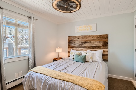 A bedroom with a wooden headboard, bed with gray and yellow linens, a window with gray curtains, a ceiling fan, and a wall sign reading "You are my sunshine.