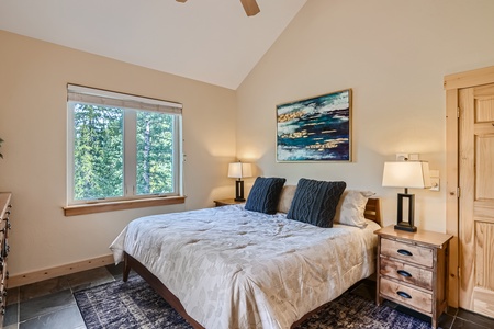 A bedroom with a large bed, two bedside tables with lamps, a window with a forest view, and a painting on the wall above the bed.