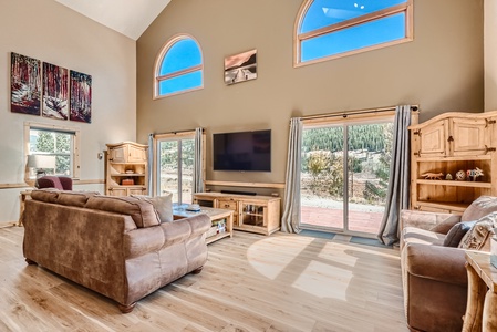 Spacious living room with high ceilings, a large flat-screen TV, brown sofa and armchair, wooden furniture, and large windows with scenic outdoor views. Light wooden floors and wall art.