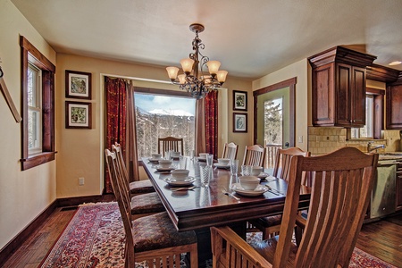 A dining room with a wooden table set for eight. Large windows show a scenic view of snow-covered mountains. The room features wooden chairs, a chandelier, and framed artwork on the walls.