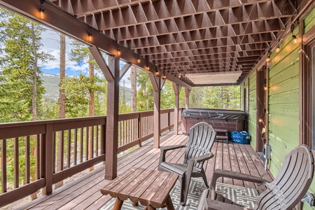 Covered wooden porch with chairs, a hot tub, and string lights, overlooking a forested area with tall trees.