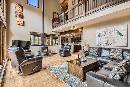 A spacious living room with tall ceilings, large windows, leather furniture, a flat-screen TV, a fireplace, and an upstairs loft area. The room has wooden floors and a modern decor.