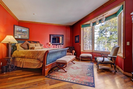 A cozy bedroom with red walls features a large bed, a wooden bench, armchairs, and a rug on a wooden floor. A window allows natural light to filter in, showcasing an outdoor view.