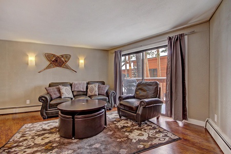 A cozy living room features a leather sofa and armchair, a round coffee table, and wall-mounted lights. A sliding glass door on the right opens to a balcony with a view of trees and a wooden building.