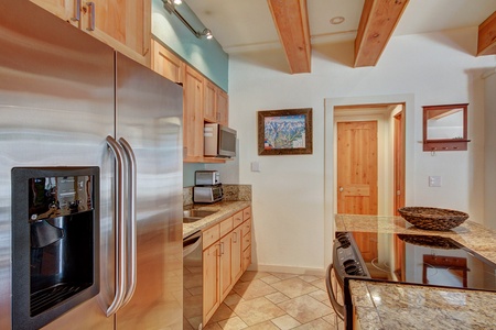 Modern kitchen with stainless steel appliances, wooden cabinets, tiled flooring, and exposed wooden beams. Features an island with a granite countertop and a colorful painting on the wall.