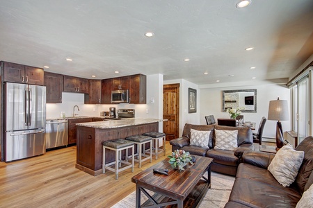 Modern kitchen and living area with wood flooring, dark wood cabinets, stainless steel appliances, a kitchen island with stools, brown leather sofas, a wooden coffee table, and various decor items.