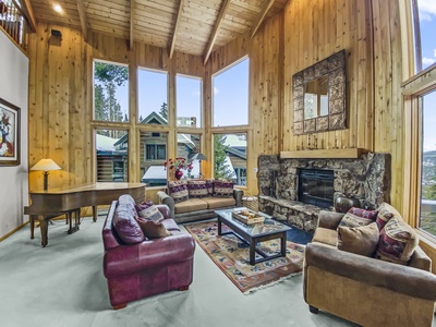 Spacious living room with large windows, wooden walls, vaulted ceiling, stone fireplace, sofas, a piano, glass coffee table, and a patterned area rug. Snowy landscape visible through the windows.