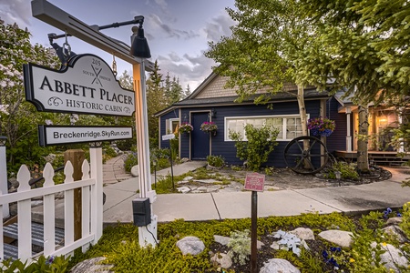 A historic cabin with a sign that reads "Abbett Placer Historic Cabin" is surrounded by greenery and flowers. A white picket fence and a stone-paved path lead to the entrance.