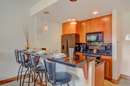 A modern kitchen featuring wooden cabinets, black granite countertops, stainless steel appliances, a breakfast bar with four stools, and decorative pendant lighting.