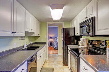 A modern, galley-style kitchen with black appliances, white cabinets, and dark countertops. There is a view into a bedroom with a nightstand and lamp.