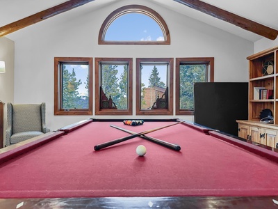 A red-felted billiard table with a white cue ball and arranged pool balls is inside a room with wooden beams and large windows overlooking trees. There are cushioned chairs and wooden shelves to the sides.