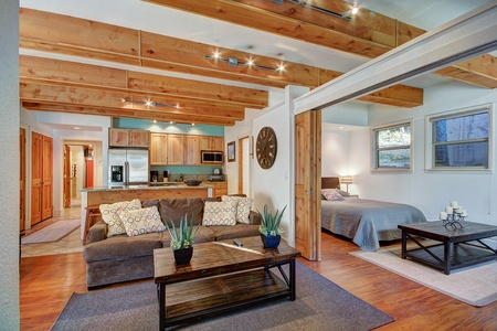 Open-plan living area with wooden ceiling beams, a kitchen with stainless steel appliances, a gray couch, a wooden coffee table, and an adjacent bedroom. The space is decorated with simple, modern furniture.