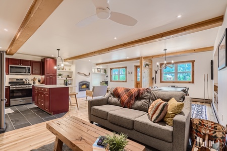 A modern open-concept living room and kitchen with a gray sofa, wooden beams, red kitchen cabinets, a fireplace, and a large rug. The space features bright lighting and a mix of contemporary and rustic decor.