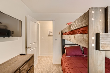 A small bedroom with a wooden bunk bed, a wall-mounted TV, a dresser with a remote on top, an open door revealing a hallway, and a painting on the wall. The bed is made with red bedding.