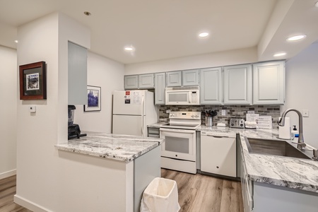 Modern kitchen with white cabinets, marble countertops, white appliances, and light wood flooring. The space includes a sink, stove, microwave, refrigerator, and small appliances.