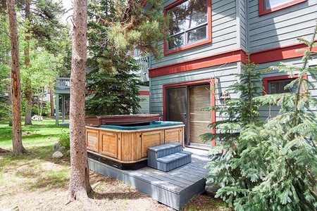 Backyard of a house with a hot tub on a wooden deck, surrounded by trees and greenery. The house has gray siding with red trim.