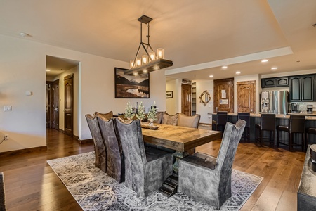 A dining room with a large wooden table, eight upholstered chairs, a modern chandelier, and an area rug. The room opens to a kitchen with a breakfast bar. Walls and floors are wooden. A painting hangs on the wall.