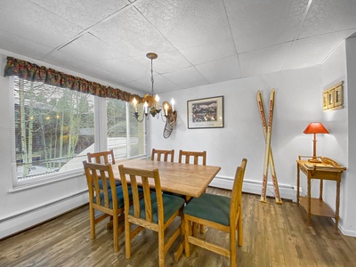 Dining room with wooden table and chairs, a chandelier, large windows with green curtains, wall decorations including skis, and a small table with a lamp in the corner.