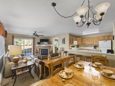 A cozy, well-lit living room and dining area with wooden furniture, a chandelier, a ceiling fan, and an open kitchen in the background. The space includes a fireplace, flat-screen TV, and a large window.