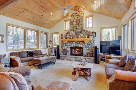 A cozy living room with a stone fireplace, wooden ceiling, large windows, beige sofas, a TV, and a coffee table. The room has a woodland view outside.