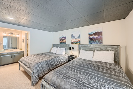 A bedroom with two double beds, grey bedding, and patterned designs. There are three landscape paintings on the wall and an adjacent bathroom with a double sink and vanity with a mirror.