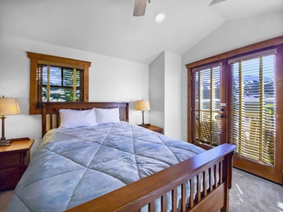 A bedroom with a large wooden bed, light blue comforter, two side tables with lamps, wooden blinds, and a glass door leading outside.