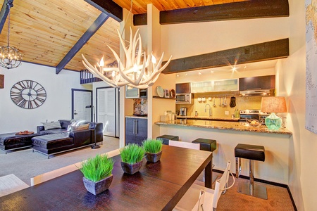 A modern living room and kitchen with wood ceilings, a chandelier, black leather furniture, a clock on the wall, a kitchen island with bar stools, and a dining table with potted plants.