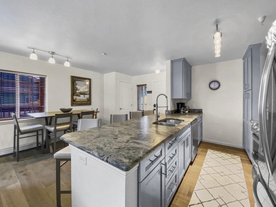 Modern kitchen with a large island, light blue cabinets, hanging light fixtures, stainless steel appliances, a dining table with chairs, and a rug on the wooden floor. Wall clock and artwork are visible.