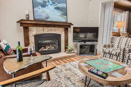 A cozy living room with a fireplace, TV, Monopoly game on the coffee table, a bottle of wine with two glasses on a side table, and armchairs with a patterned design.