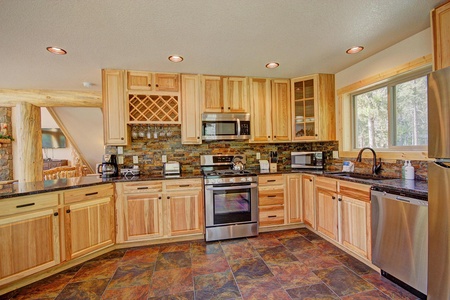 A modern kitchen with wooden cabinets, stainless steel appliances, a stove, microwave, dishwasher, and a tiled backsplash. A window provides natural light and there are wooden beams visible in the background.