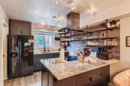 Modern kitchen with black appliances, granite countertops, and open shelving. Central island features an induction cooktop and utensils holder. Track lighting illuminates the space.