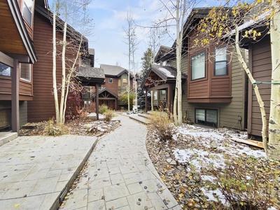 A narrow stone pathway meanders through a courtyard of brown wooden townhouses with some snow scattered on the ground. Leafless trees line the sides, indicating early winter.