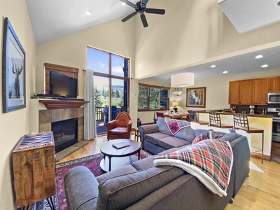 A modern living room with a gray sofa, armchair, TV above a fireplace, and large windows. The space is open to a kitchen with wooden cabinets, bar seating, and a dining area.