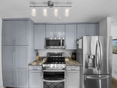 Modern kitchen with light gray cabinets, stainless steel appliances including a refrigerator, microwave, and stove, granite countertops, and a ceiling light fixture with five bulbs.