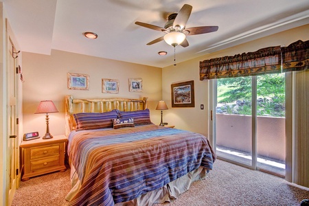 A well-lit bedroom with a ceiling fan features a bed with striped bedding, two bedside tables with lamps, framed pictures on the walls, and a sliding glass door leading to an outdoor space.