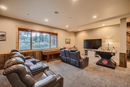 A home theater room features leather recliners, a shuffleboard table, a flat-screen TV on a media stand, an arcade hockey table, and a large window offering views of an outdoor wooded area.