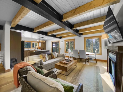 A cozy living room with a modern design, featuring exposed wooden beams, a large couch, a fireplace, and a wall-mounted TV. The room opens to a kitchen and dining area with large windows.