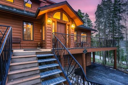 Exterior of a log house at sunset with illuminated lights, a steep staircase, and metal railings leading to a wood front door. Trees and a road are visible in the background.