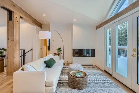 A modern living room featuring a white sofa, a round wicker coffee table, a large white floor lamp, and a fireplace. The room has wooden beams, a patterned rug, and large windows with a view outside.