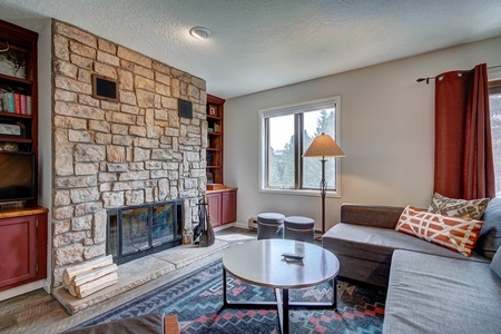 A cozy living room with a stone fireplace, a sectional sofa, a round coffee table, a standing lamp, and built-in shelves. A window and red curtains flank the fireplace.