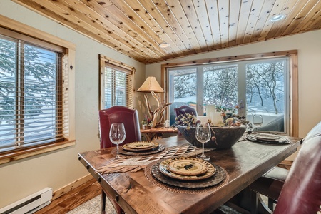 A rustic dining room with a wooden table set for four, featuring wine glasses and decorative plates. The room has wood-paneled ceilings and large windows with views of snow-covered trees outside.