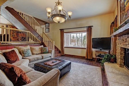 A cozy living room with a beige sectional sofa, a brown ottoman, a stone fireplace, a flat-screen TV, and large windows by a staircase. The floor is hardwood with a rug in the center.