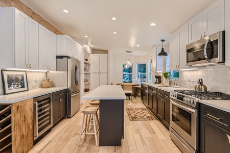 Modern kitchen with white and black cabinetry, stainless steel appliances, an island with stools, and a small dining area with a window. Light wood flooring and recessed lighting complete the space.