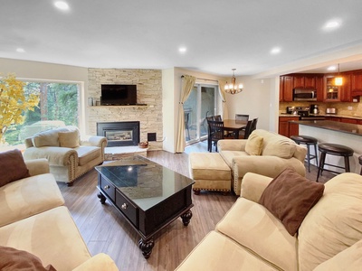 A cozy living room with beige sofas, brown pillows, a black coffee table, and a fireplace. Dining area and kitchen with bar stools are visible in the background. Large windows allow natural light.