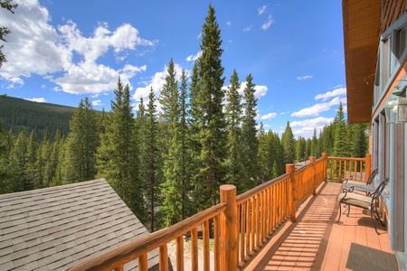 A cabin balcony with wooden railing overlooks a dense forest of tall pine trees under a clear blue sky with scattered clouds.