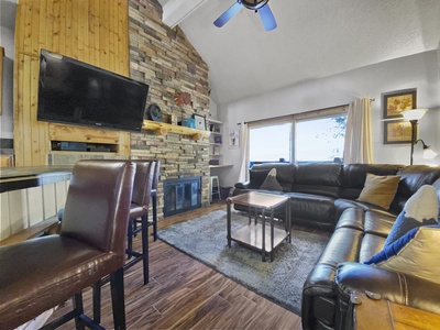 A living room with a stone fireplace, wall-mounted TV, brown leather couches, a wooden coffee table, and large window with curtains. A ceiling fan is visible, and the floor is made of wood.