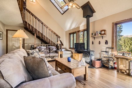 A cozy living room with wooden furniture, a fireplace, and a TV. Stairs lead to an upper level, and sunlight streams through skylights and a window with a view of trees.